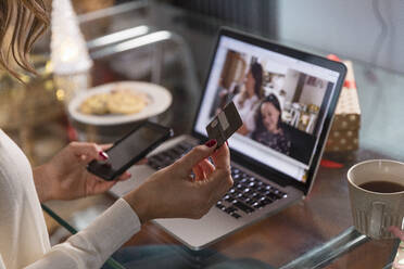 Frau mit Kreditkarte und Smartphone bei einem Videogespräch mit Freunden über einen Laptop zu Hause - EIF04117