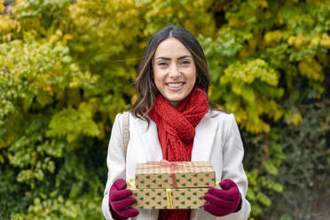 Glückliche Frau mit rotem Schal steht mit Weihnachtsgeschenken im Park - EIF04146