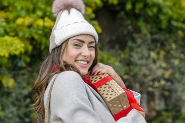 Schöne glückliche Frau mit Strickmütze umarmt Weihnachtsgeschenk im Park - EIF04153