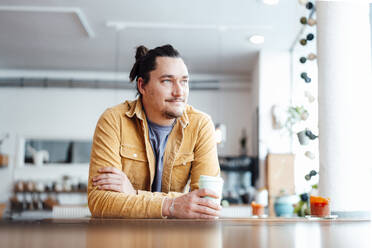 Smiling man with disposable coffee cup sitting at table in cafe - JOSEF09879