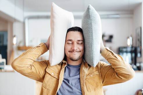 Smiling man covering ears with cushions at cafe - JOSEF09884