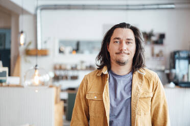 Smiling hipster man with long hair standing at cafe - JOSEF09886