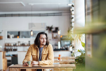 Hipster-Mann mit Kaffeetasse, der sich in einem Café auf einen Tisch stützt - JOSEF09891