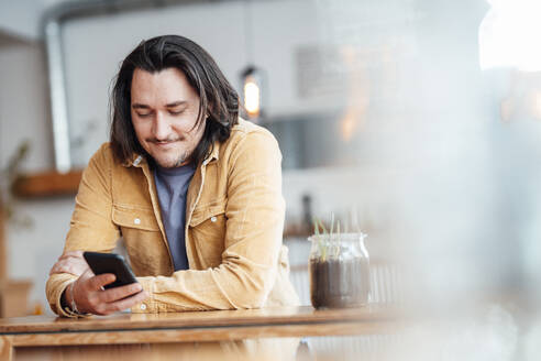 Lächelnder Mann mit langen Haaren, der sich auf einen Tisch stützt und ein Smartphone im Café benutzt - JOSEF09895