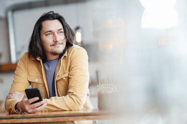 Lächelnder Mann mit Mobiltelefon, der sich in einem Café auf einen Tisch stützt - JOSEF09897