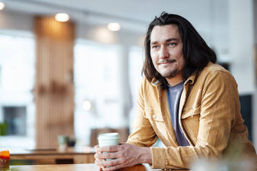 Lächelnder Hipster-Mann mit Kaffeetasse, der sich in einem Café auf einen Tisch stützt - JOSEF09904