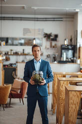 Mature businessman with plant in glass container standing at cafe - JOSEF09927