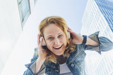 Fröhliche Frau hört Musik über drahtlose Kopfhörer inmitten von Gebäuden - IHF00844