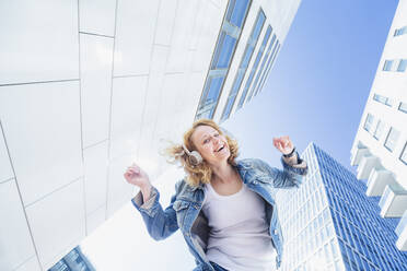 Happy woman enjoying music through wireless headphones standing amidst buildings - IHF00845