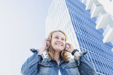 Smiling woman listening music through wireless headphones standing in front of building - IHF00846