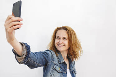 Lächelnde Frau spricht Selfie durch Smartphone gegen weißen Hintergrund - IHF00851