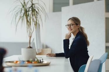 Smiling businesswoman sitting with hand on chin at desk - JOSEF09959
