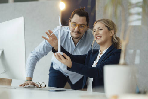 Smiling businesswoman holding wind turbine model discussing with colleague in office - JOSEF09967