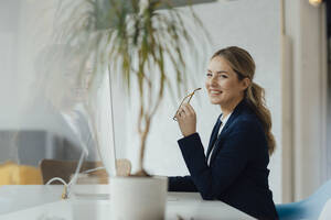 Lächelnde Geschäftsfrau mit Brille sitzt am Schreibtisch im Büro - JOSEF09971