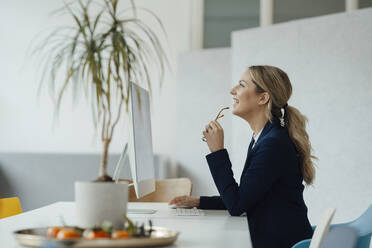 Happy businesswoman holding eyeglasses sitting at computer desk - JOSEF09973