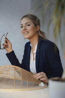 Lächelnder Architekt mit hölzernem Modell in Form eines Blattes am Schreibtisch - JOSEF09978