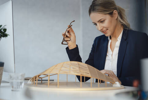 Lächelnde Geschäftsfrau, die ein Modell in Form eines Holzblattes am Schreibtisch analysiert - JOSEF09979