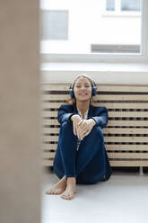 Smiling businesswoman listening music through wireless headphones leaning on radiator in office - JOSEF09986