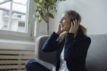 Happy businesswoman enjoying music listening through wireless headphones in office - JOSEF09995