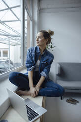 Smiling freelancer with laptop sitting cross-legged on widow sill at home - JOSEF10006