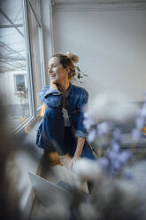 Happy businesswoman with laptop sitting on widow sill at home - JOSEF10007