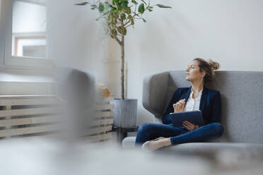 Thoughtful businesswoman holding tablet PC sitting on sofa in office - JOSEF10028