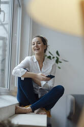 Happy woman holding smart phone sitting on widow sill at home - JOSEF10037