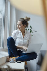 Happy businesswoman with laptop sitting on widow sill in office - JOSEF10038