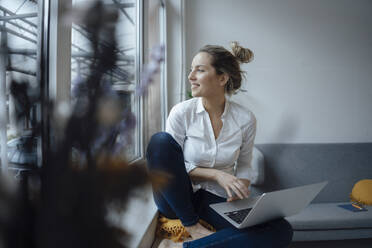 Smiling businesswoman with laptop sitting on widow sill in office - JOSEF10039
