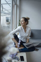 Happy businesswoman holding laptop sitting on widow sill in office - JOSEF10040