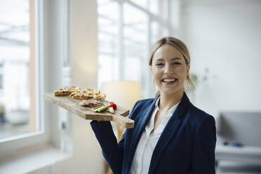 Smiling beautiful businesswoman with sandwich on cutting board in office - JOSEF10049