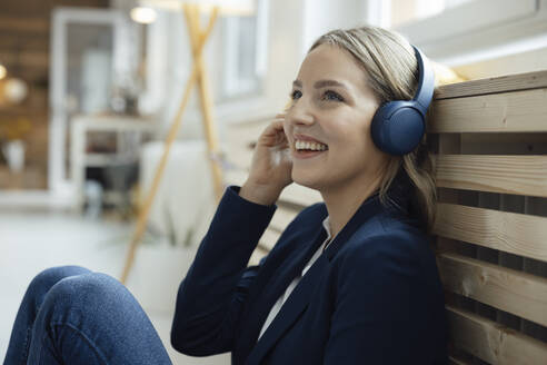 Glückliche Geschäftsfrau, die über drahtlose Kopfhörer Musik hört und sich im Büro an einen Heizkörper lehnt - JOSEF10055