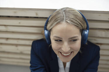 Lächelnde Geschäftsfrau, die über drahtlose Kopfhörer Musik hört und sich im Büro an einen Heizkörper lehnt - JOSEF10056