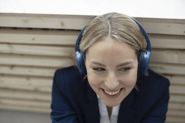 Smiling businesswoman enjoying music listening through wireless headphones leaning on radiator in office - JOSEF10056