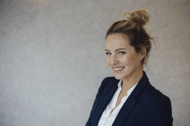 Happy businesswoman with blond hair bun in front of gray wall - JOSEF10068