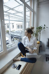 Happy businesswoman with laptop sitting on window sill at home - JOSEF10077