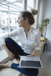 Smiling freelancer with laptop sitting on window sill at home - JOSEF10078