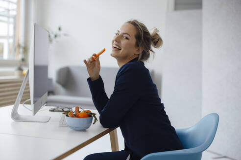Happy businesswoman holding carrot sitting at desk in office - JOSEF10090