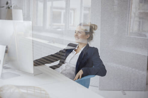 Glückliche Geschäftsfrau, die einen Desktop-PC am Schreibtisch benutzt, gesehen durch Glas - JOSEF10092