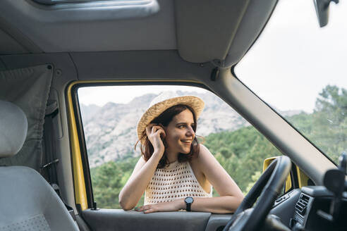 Happy woman standing with hand in hair at van window - DAMF00961