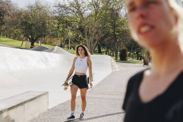 Lächelnde Frau mit Skateboard in der Nähe eines Freundes auf einer Sportrampe an einem sonnigen Tag - MRRF02148