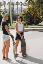 Smiling friends holding skateboards talking at sports ramp on sunny day - MRRF02153