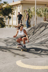 Junge Frau fährt Skateboard auf einer Sportrampe mit Freunden im Hintergrund - MRRF02175