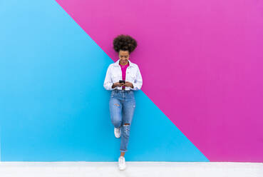 Smiling woman using smart phone standing on one leg leaning on pink and blue wall - OIPF01775