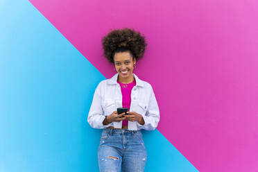 Smiling young woman with mobile phone standing in front of pink and blue wall - OIPF01776
