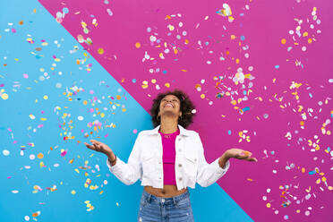 Happy woman with Afro hairstyle standing amidst confetti in front of pink and blue wall - OIPF01778