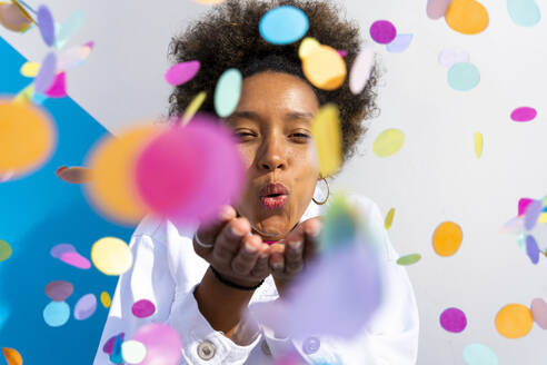 Young woman blowing multi colored confetti in front of wall on sunny day - OIPF01779