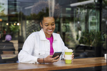 Smiling young woman with disposable coffee cup using mobile phone seen through glass - OIPF01780