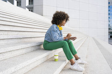 Young Afro woman with tablet PC sitting by disposable coffee cup on steps - OIPF01786