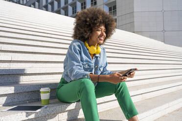 Lächelnde Afro-Frau mit drahtlosen Kopfhörern, die mit einem Mobiltelefon auf einer Treppe sitzt - OIPF01792